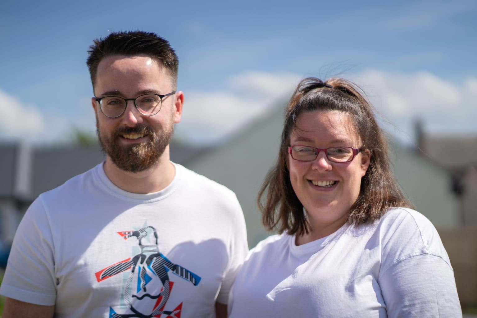 Stuart and Deborah stand smiling at the camera in white T-shirts. There is a beautiful blue sky behind them.