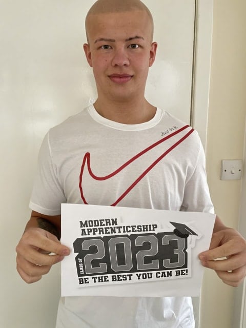 Mason is wearing a white shirt and smiling. He is holding a sign that says Modern Apprenticeships 2023.